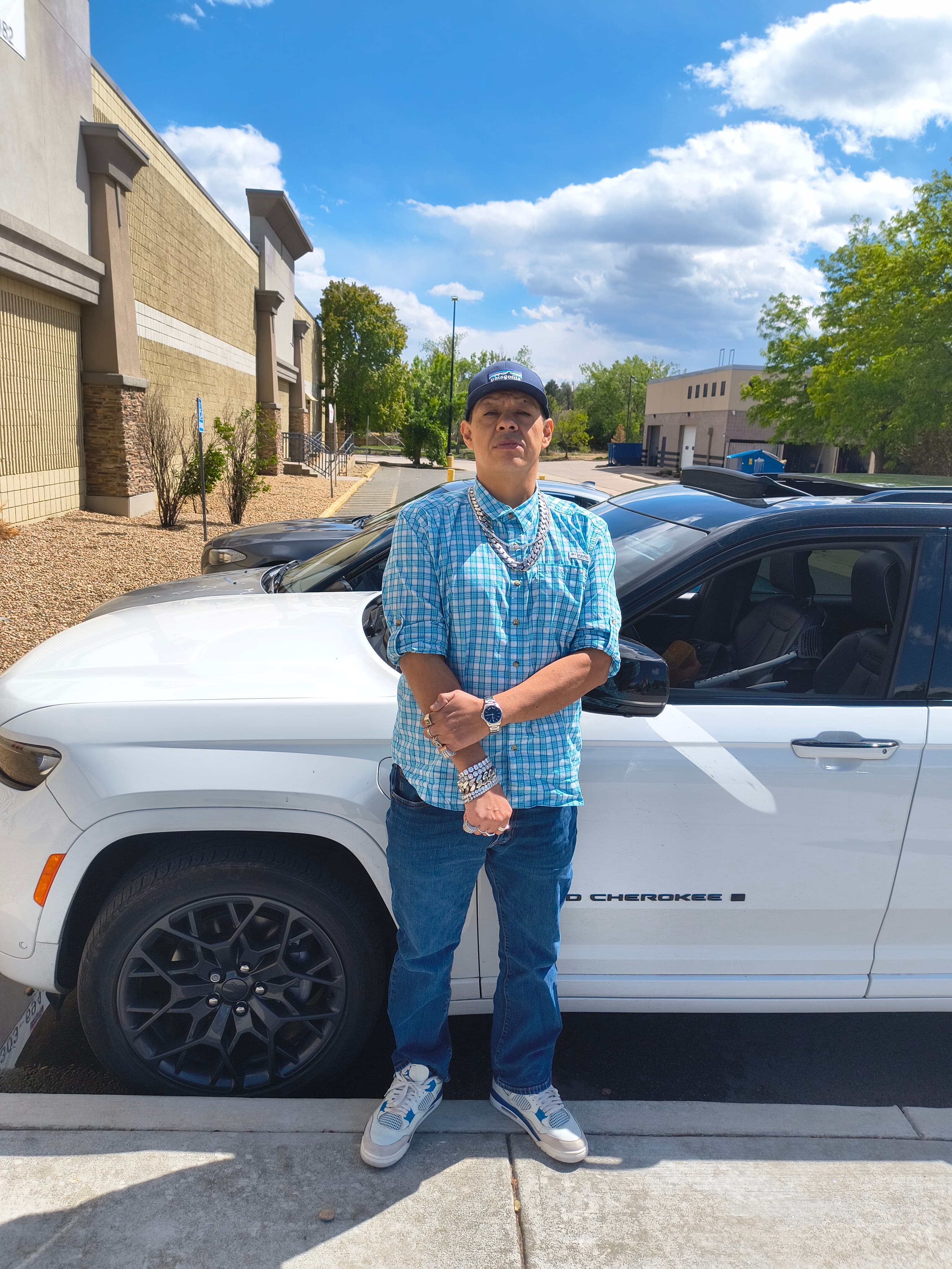 Man in blue plaid shirt and silver jewelry posing in front of a white Jeep.